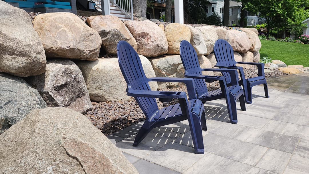 Three blue adirondack chairs on a stone patio by a rock wall in a landscaped yard.