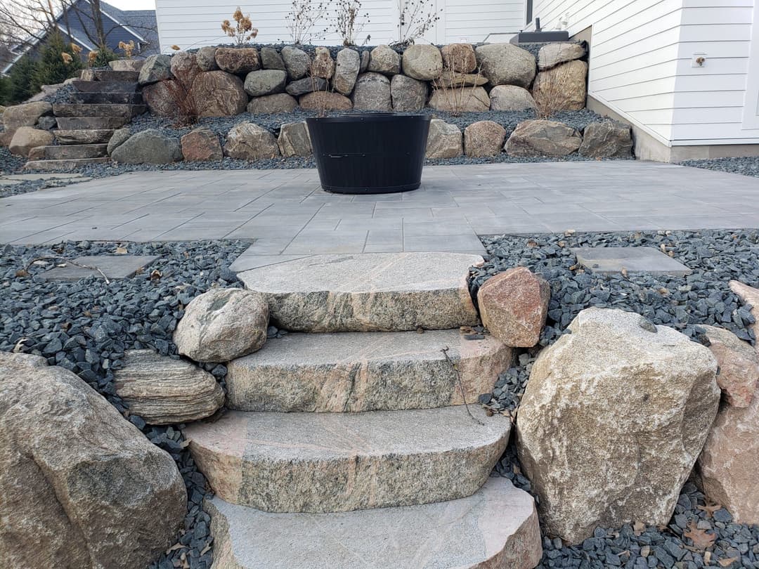 Stone steps leading to a patio with a black planter surrounded by decorative rocks and boulders.