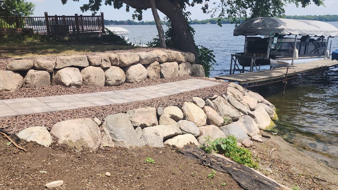 Scenic lakeside view featuring a stone path and dock with boat, surrounded by greenery.