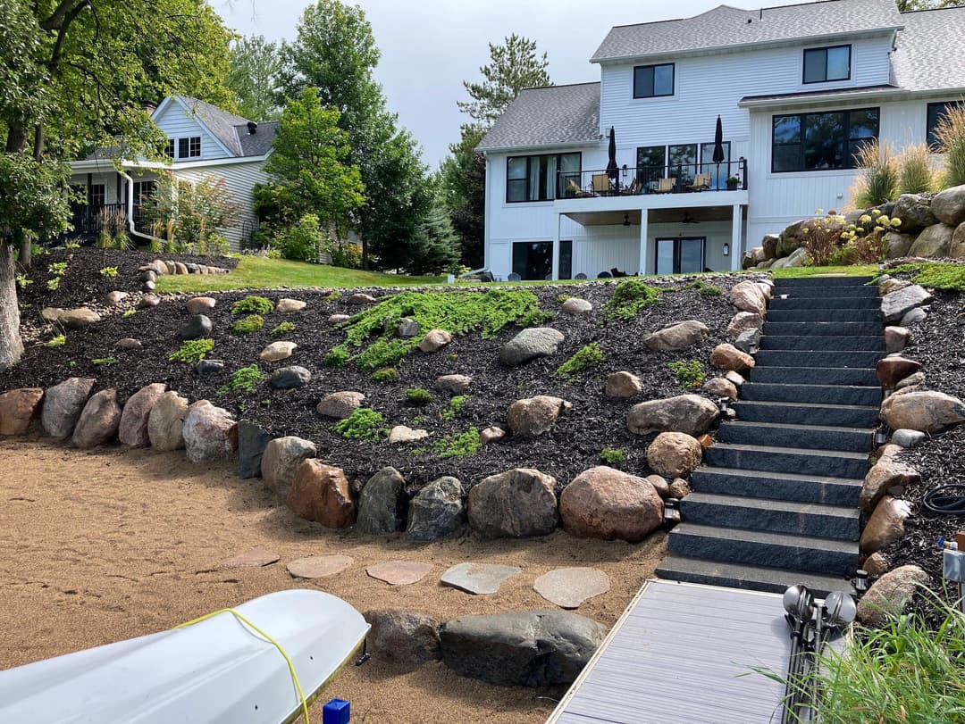 Beautiful landscaped backyard with stone steps and mulch, leading to a white house overlooking a lake.