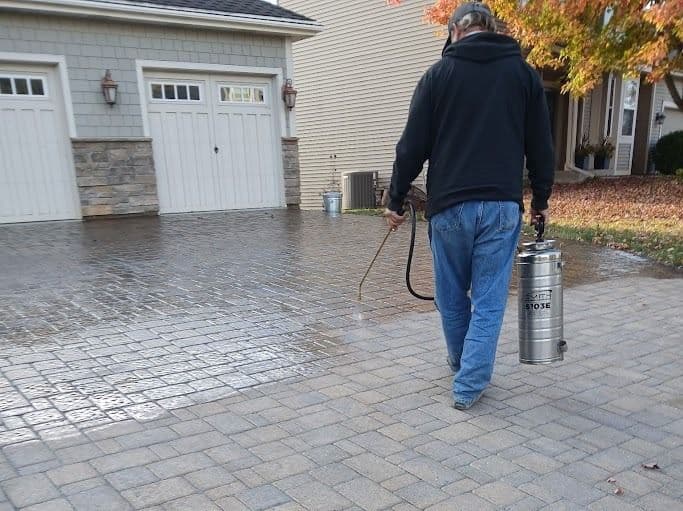 Person using a sprayer on a clean, brick driveway in a residential neighborhood.