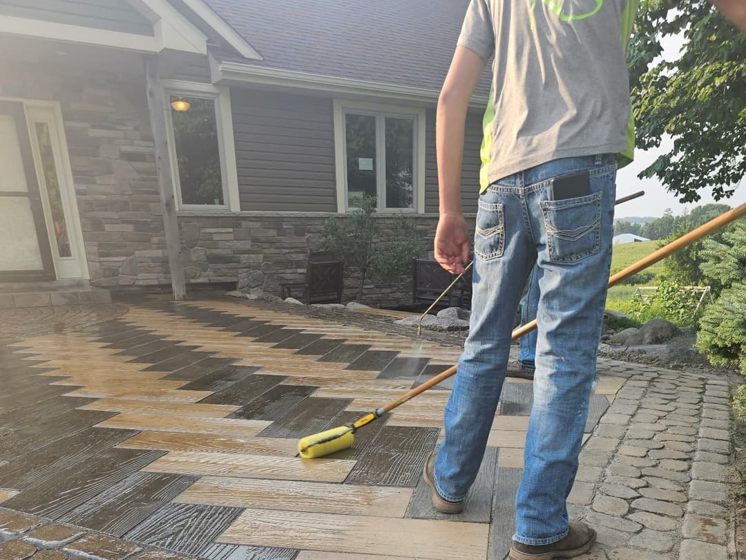 Person cleaning a patterned stone patio with a pressure washer, surrounded by greenery.