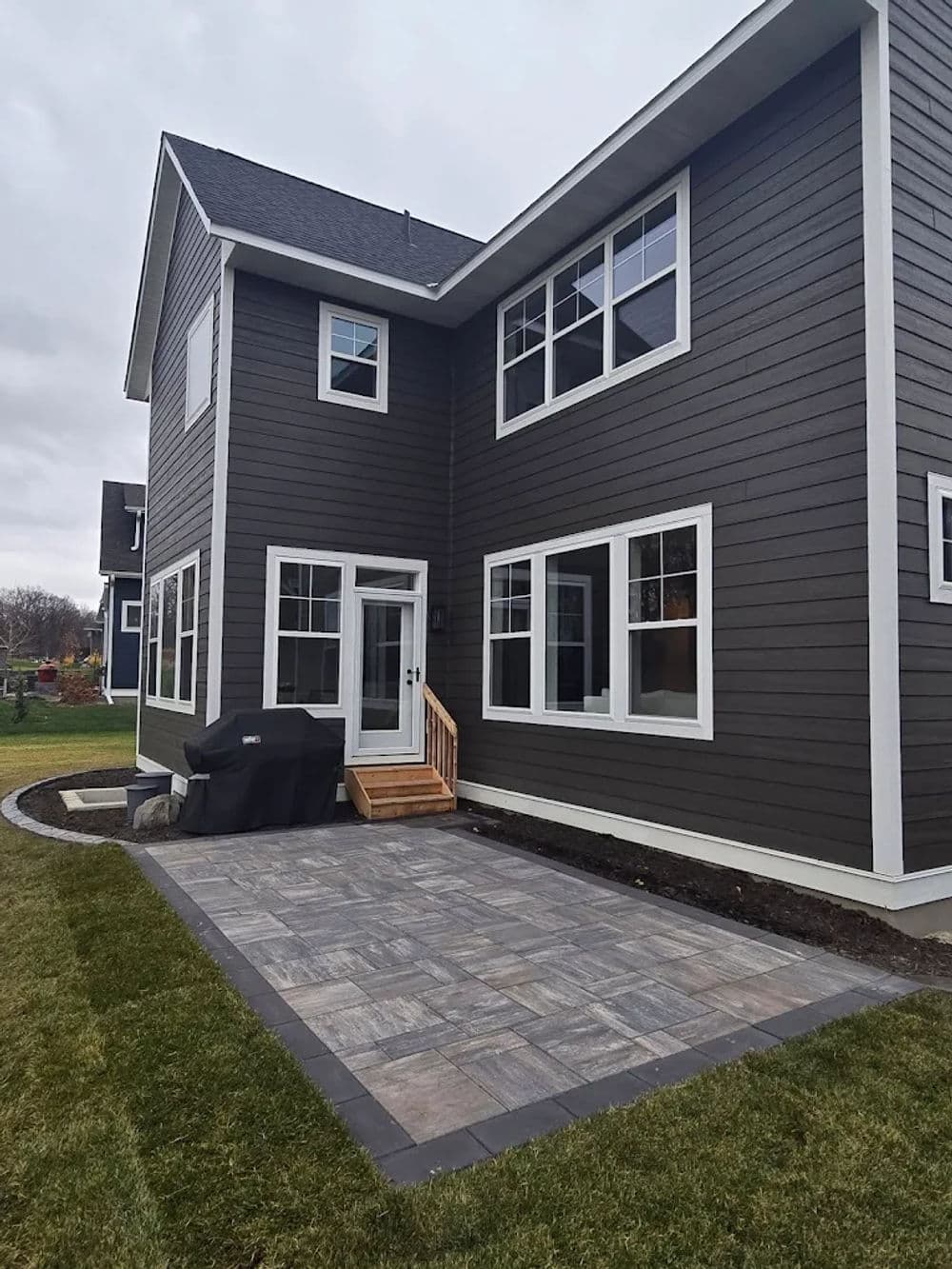 Modern home exterior with dark siding, patio, and gas grill in landscaped yard.