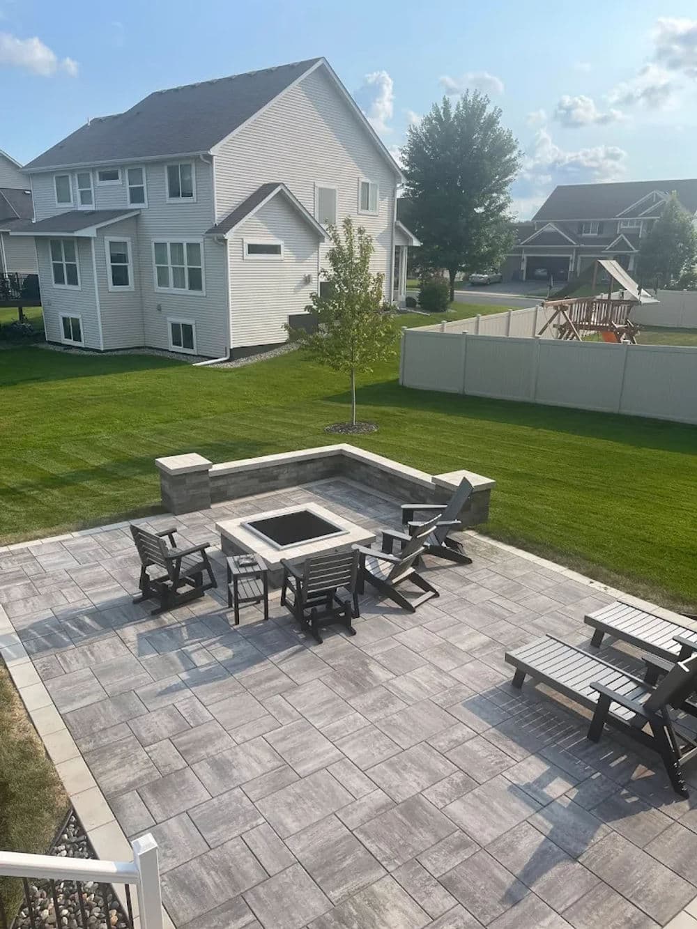 Backyard patio with fire pit, seating, and green lawn in residential area on a sunny day.
