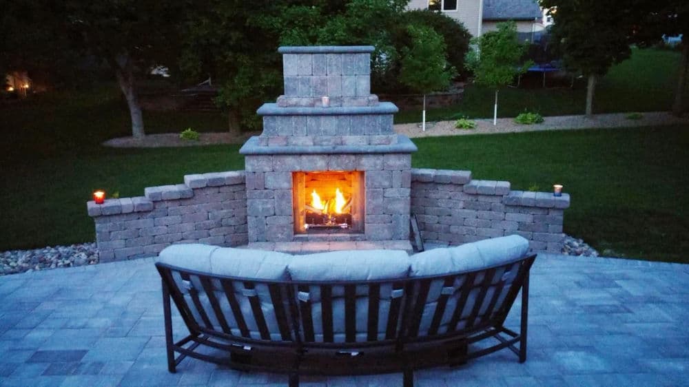 Cozy outdoor patio with a stone fireplace and seating area, illuminated at dusk.