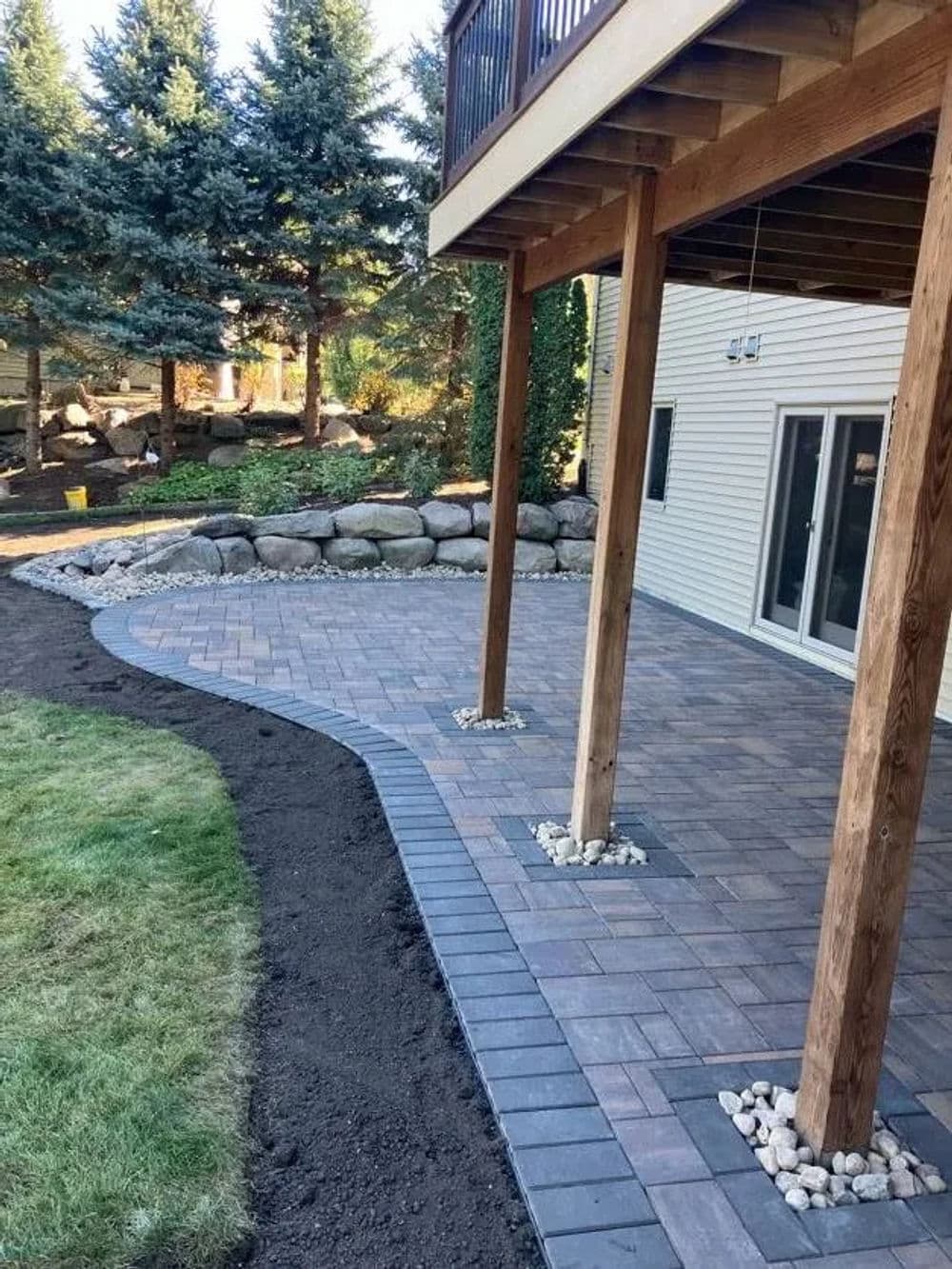 Patio area with stone pavers, gravel borders, and wooden deck in landscaped backyard.