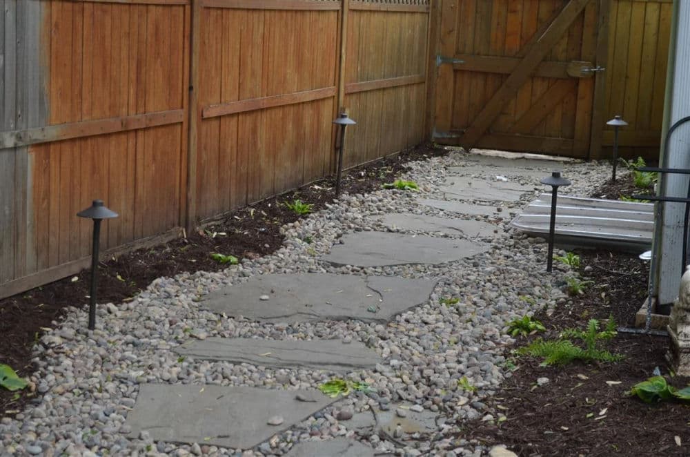 Garden pathway with stone slabs, gravel, and landscaping lights alongside wooden fence.