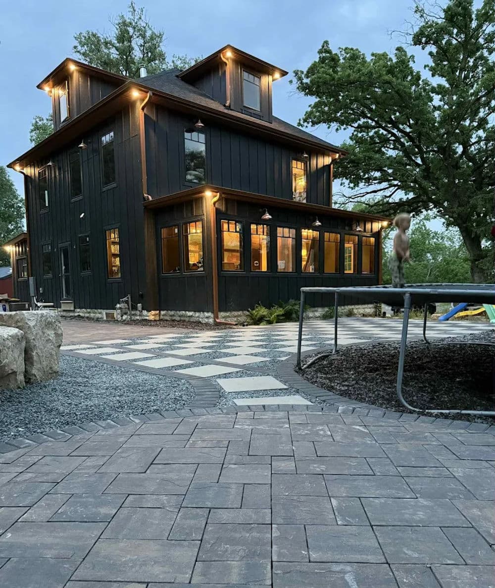 Modern black wooden house with warm lights, patio area, and stone walkway at twilight.