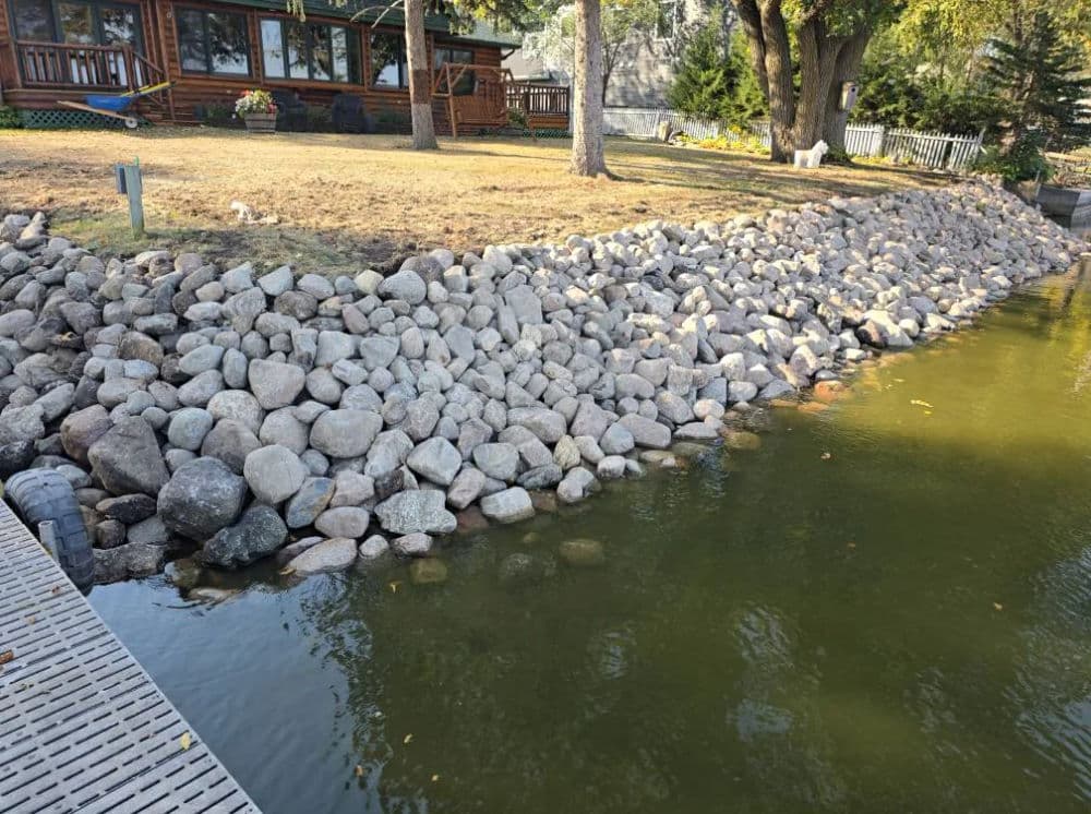 Rocky shoreline along a calm lake, with a wooden dock and house in the background.