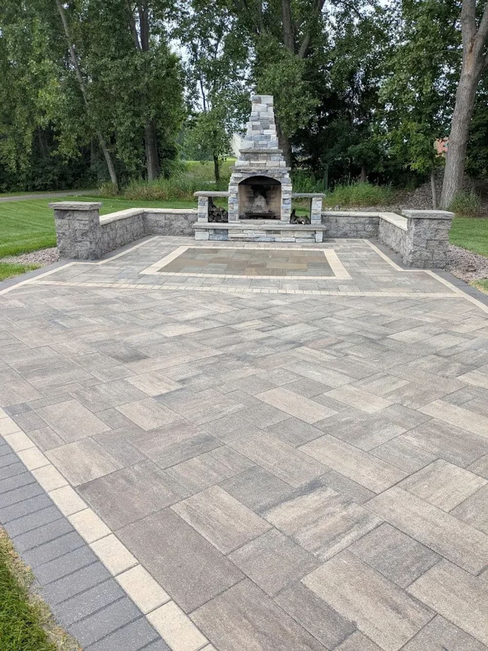 Outdoor patio with stone fireplace, surrounded by landscaped greenery and patterned pavers.