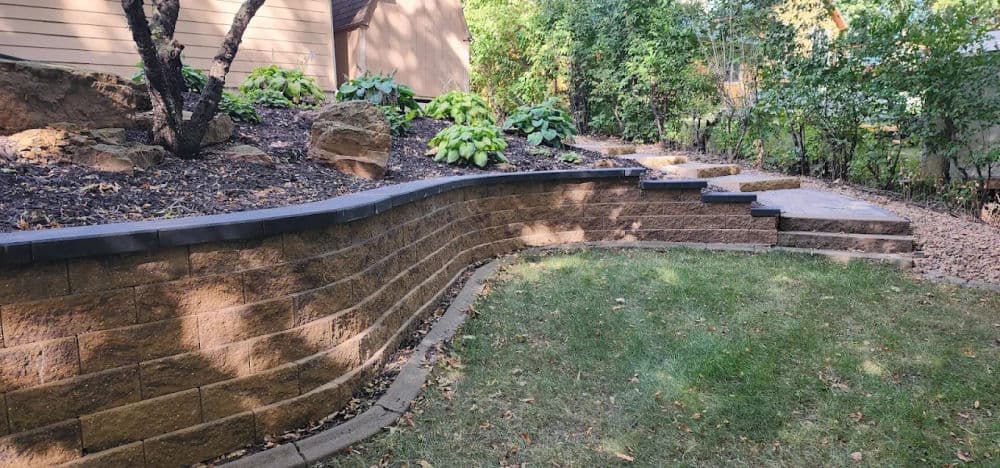 Landscaped garden with curved stone wall, steps, and lush greenery in a serene setting.