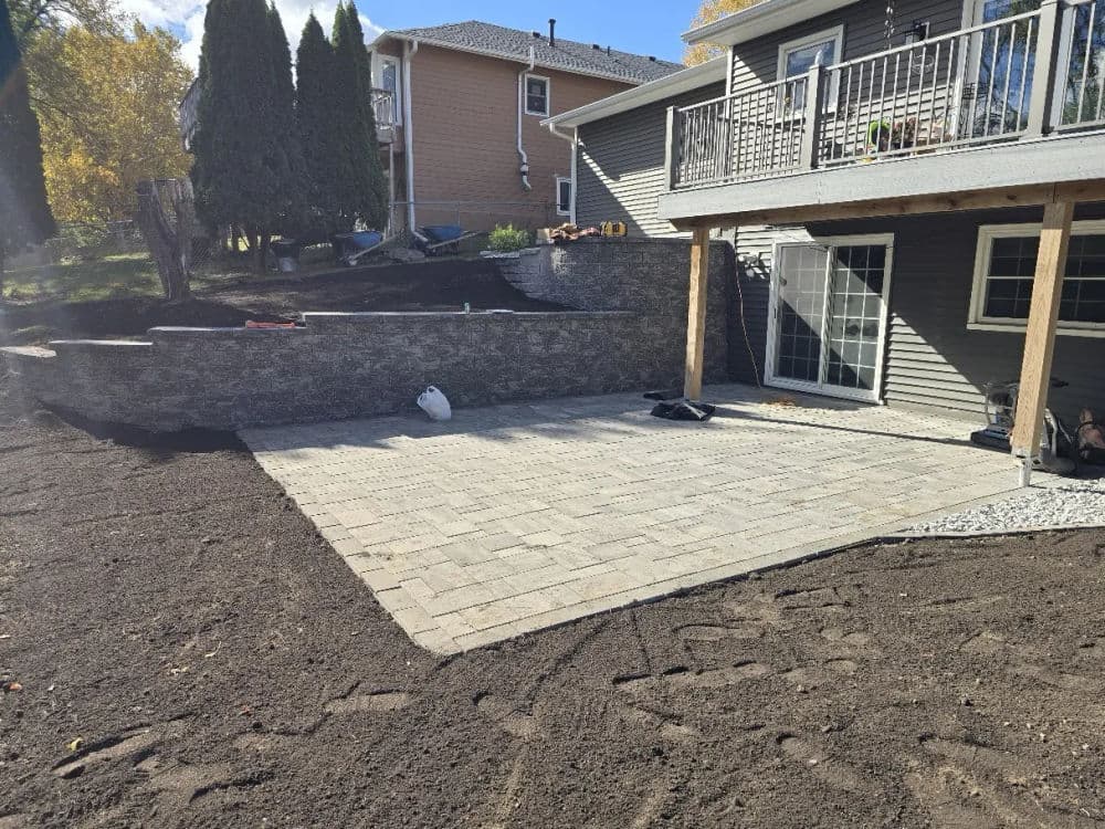 Newly installed stone patio adjacent to a house with landscaped yard and retaining wall.