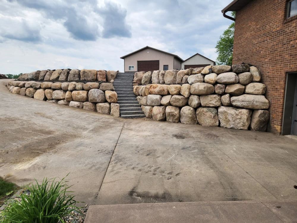 Stone retaining wall with stairs beside a driveway and two buildings in the background.