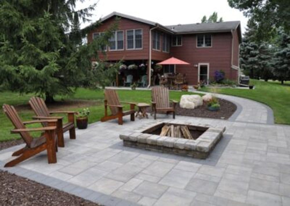 Backyard patio with stone fire pit and wooden chairs, surrounded by lush greenery and a house.