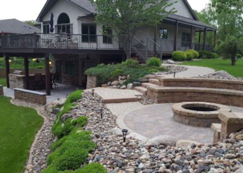 Modern home with landscaped stone patio, fire pit, and lush greenery in backyard setting.