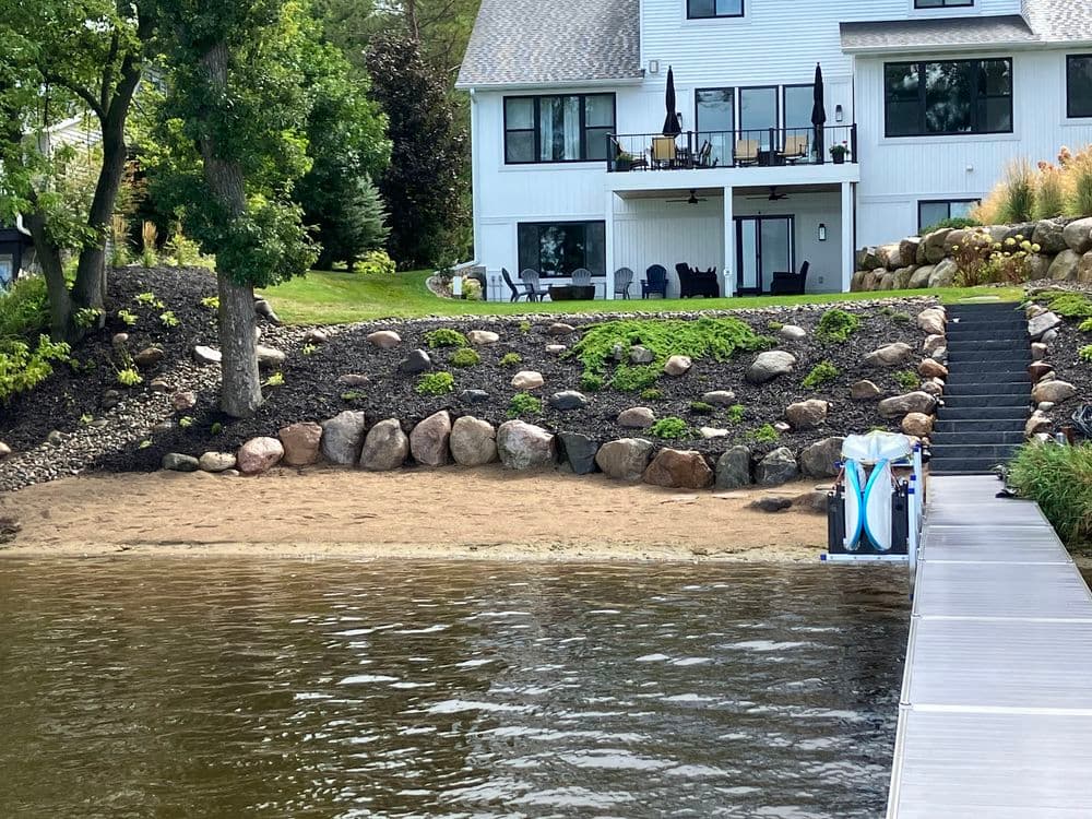 Lakeside view of a home with landscaped yard, sandy beach, and dock.
