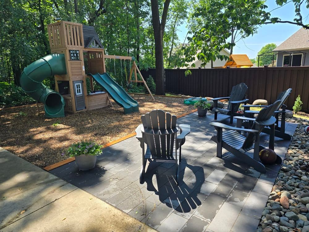 Backyard playground with slide, playhouse, and patio seating surrounded by trees.