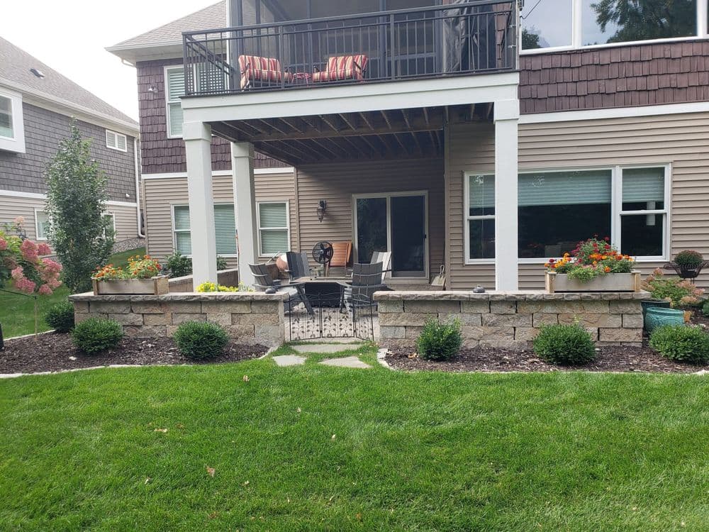 Backyard patio with seating, stone wall, and colorful flower beds surrounded by green lawn.