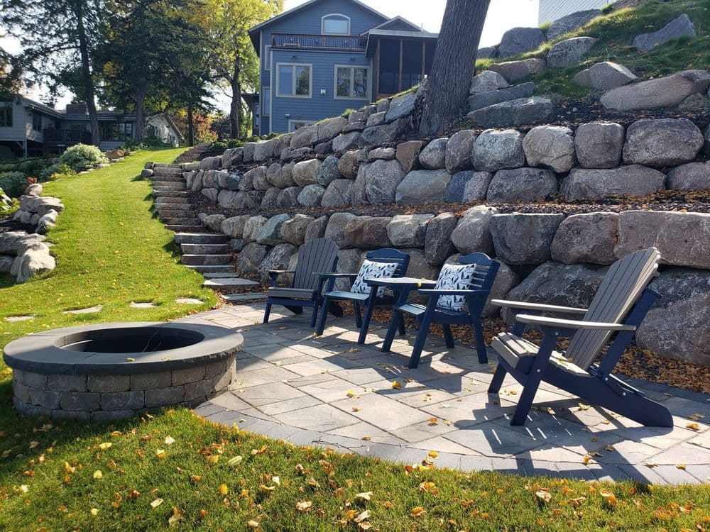 Lakeside patio with Adirondack chairs and stone fire pit surrounded by lush greenery.