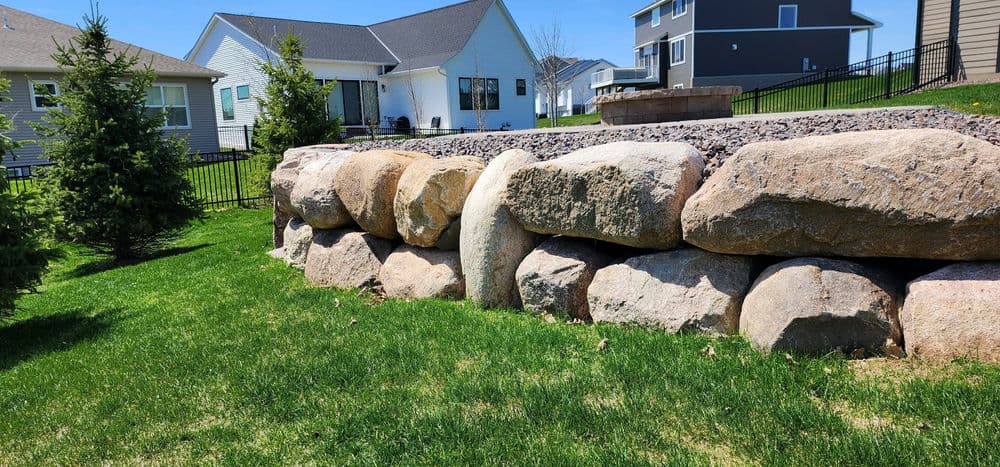 Stone retaining wall in a residential yard with green grass and modern homes in the background.