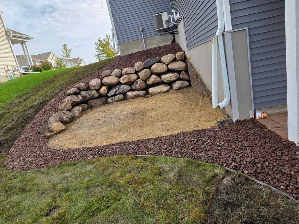 Landscaped area with decorative rocks, gravel, and a stone wall beside a house.