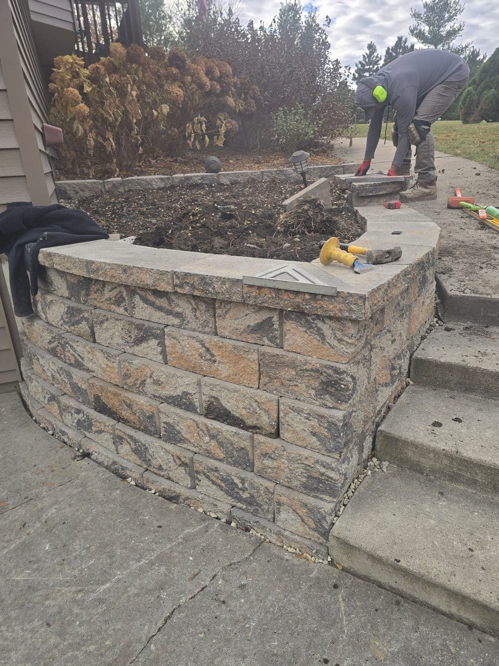 Person installing a stone garden bed with tools, surrounded by autumn foliage.