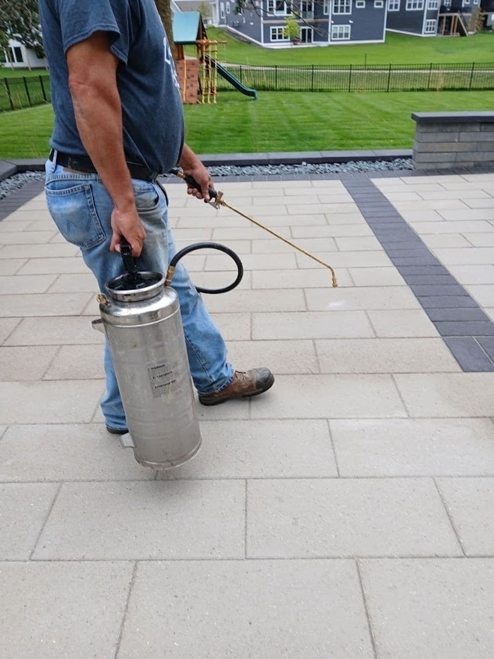 Man using a sprayer on patio pavers for maintenance in a landscaped yard.