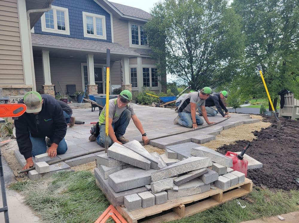 Workers installing paving stones on a residential patio, with tools and materials nearby.