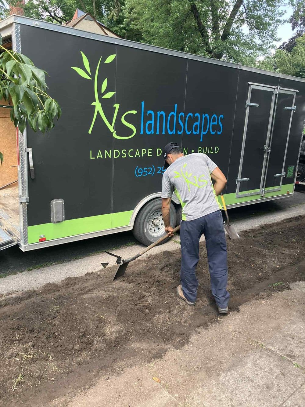 Landscape worker digging near a branded trailer for a landscaping business.
