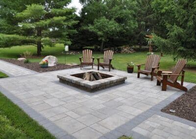 Outdoor patio with stone fire pit and wooden chairs surrounded by trees and greenery.