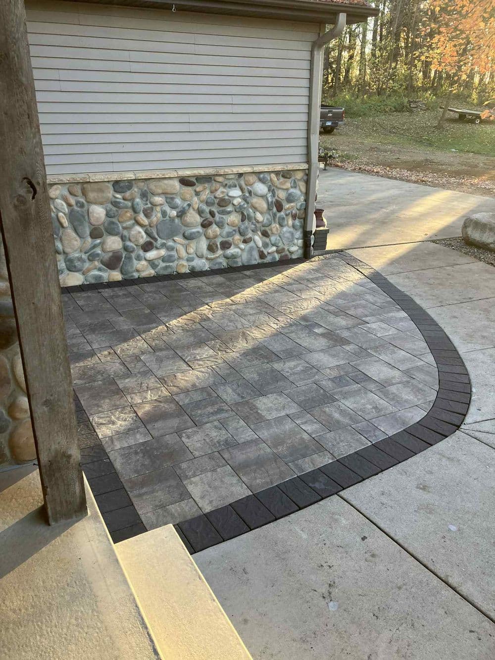 paved patio area with stone borders next to a house and driveway in a wooded setting