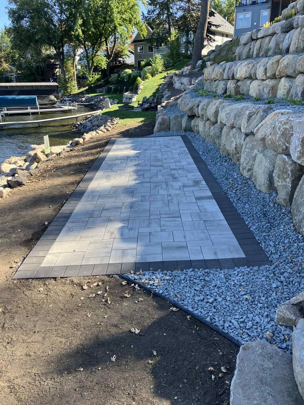 Paver patio installation by a lake with stone retaining wall and gravel landscaping.